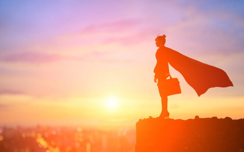 Silhouette of woman wearing a cape, sensible high heels, and a business suite standing on a cliff edge at sunrise holding a briefcase